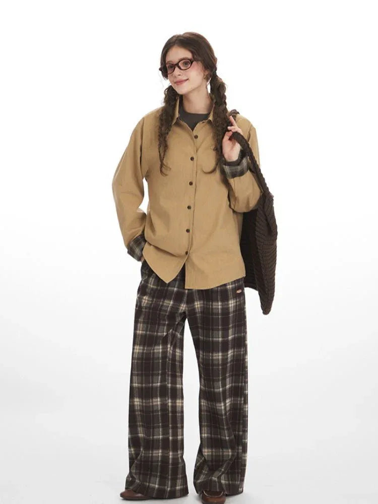 Full body shot of a woman in a tan button-up shirt and brown plaid wide-leg pants, carrying a brown knit bag.
