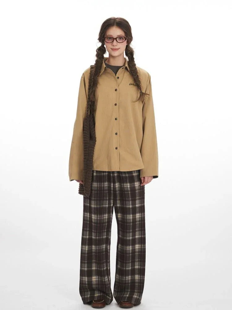 Woman wearing a tan button-up shirt with embroidered logo and plaid cuffs, paired with brown plaid wide-leg pants.