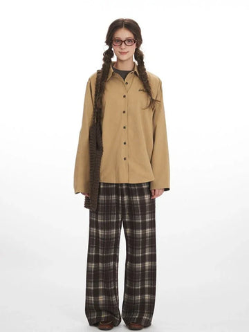Woman wearing a tan button-up shirt with embroidered logo and plaid cuffs, paired with brown plaid wide-leg pants.