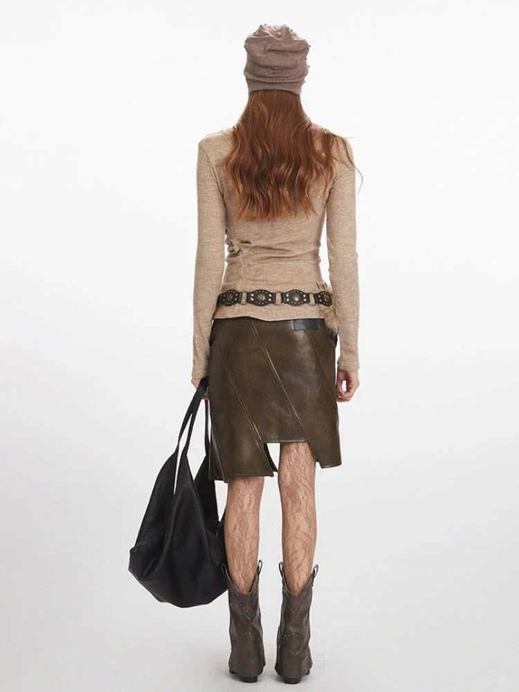 Woman wearing a long-sleeved tan knit top with draped front detail, a brown leather-look asymmetrical skirt with lace-up detail, a fur-embellished belt, brown cowboy boots, a tan knit beanie with embellishments, and carrying a black oversized tote bag.