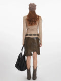 Back view of a woman wearing a long-sleeved tan knit top, a brown leather-look asymmetrical skirt, a decorative belt, brown cowboy boots, and a tan knit beanie.
