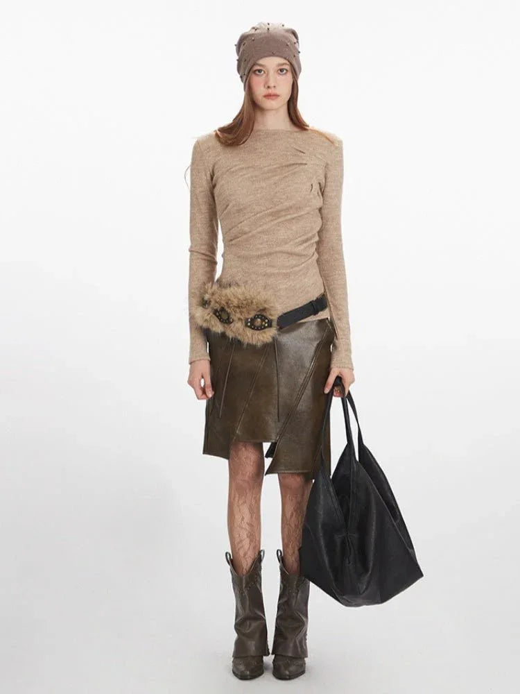 Woman wearing a long-sleeved tan knit top with draped front detail, a brown leather-look asymmetrical skirt with lace-up detail, a fur-embellished belt, brown cowboy boots, a tan knit beanie with embellishments, and carrying a black oversized tote bag.