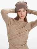 Woman in a tan knit top with draped detailing on the chest and a jeweled beanie.