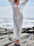 Woman wearing a white crochet maxi cover-up dress with long sleeves and a deep V-neckline over a white bikini on a rocky beach.