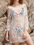 Woman wearing a white crochet knit tunic over a patterned bikini, standing by rocks at the beach.