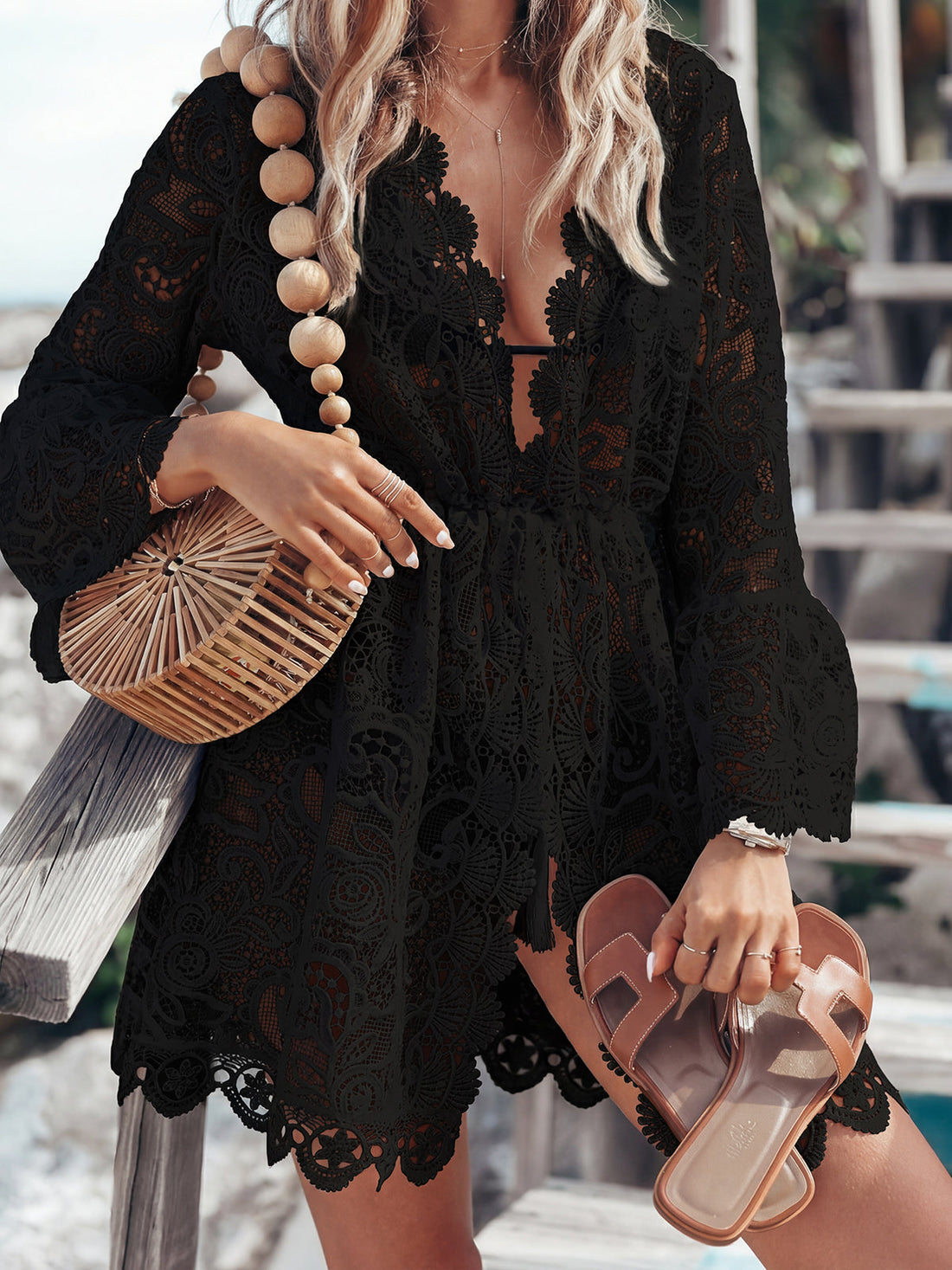 Woman wearing a white lace cover-up with bell sleeves and a deep V-neckline, holding a straw bag and sandals.