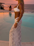 Side view of a woman in a white bikini and white lace maxi skirt with cut-outs, by a pool.