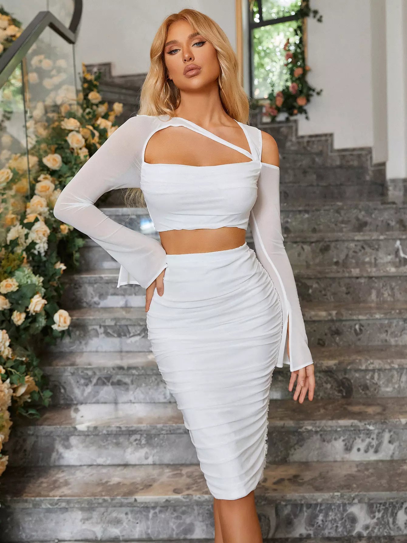 Woman in a white two-piece set with an asymmetrical crop top and ruched midi skirt, standing on a marble staircase.