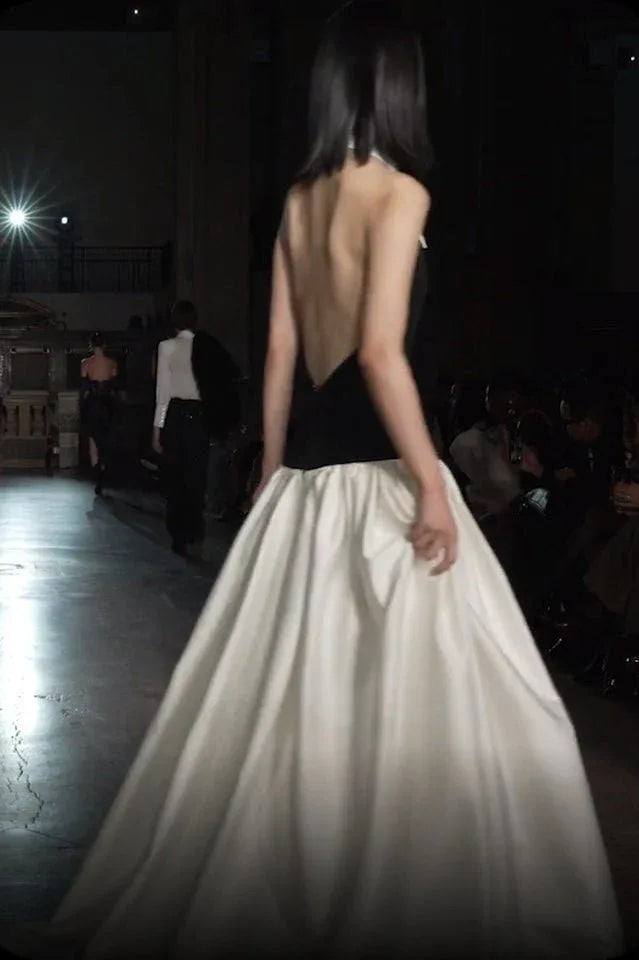 Model on a runway wearing a black halter top dress with a white pointed collar and a full ivory skirt, seen from the back.