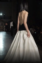 Model on a runway wearing a black halter top dress with a white pointed collar and a full ivory skirt, seen from the back.