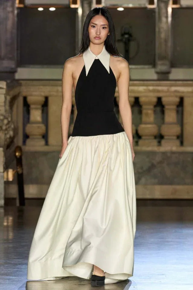 Model on a runway wearing a black halter top dress with a white pointed collar and a full ivory skirt.