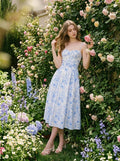 Woman wearing a blue and white floral print midi dress with spaghetti straps, standing near a rose bush in a garden.