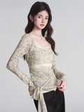Woman wearing a light-colored, long-sleeved sheer mesh top with a floral-like print and a tie detail at the waist.