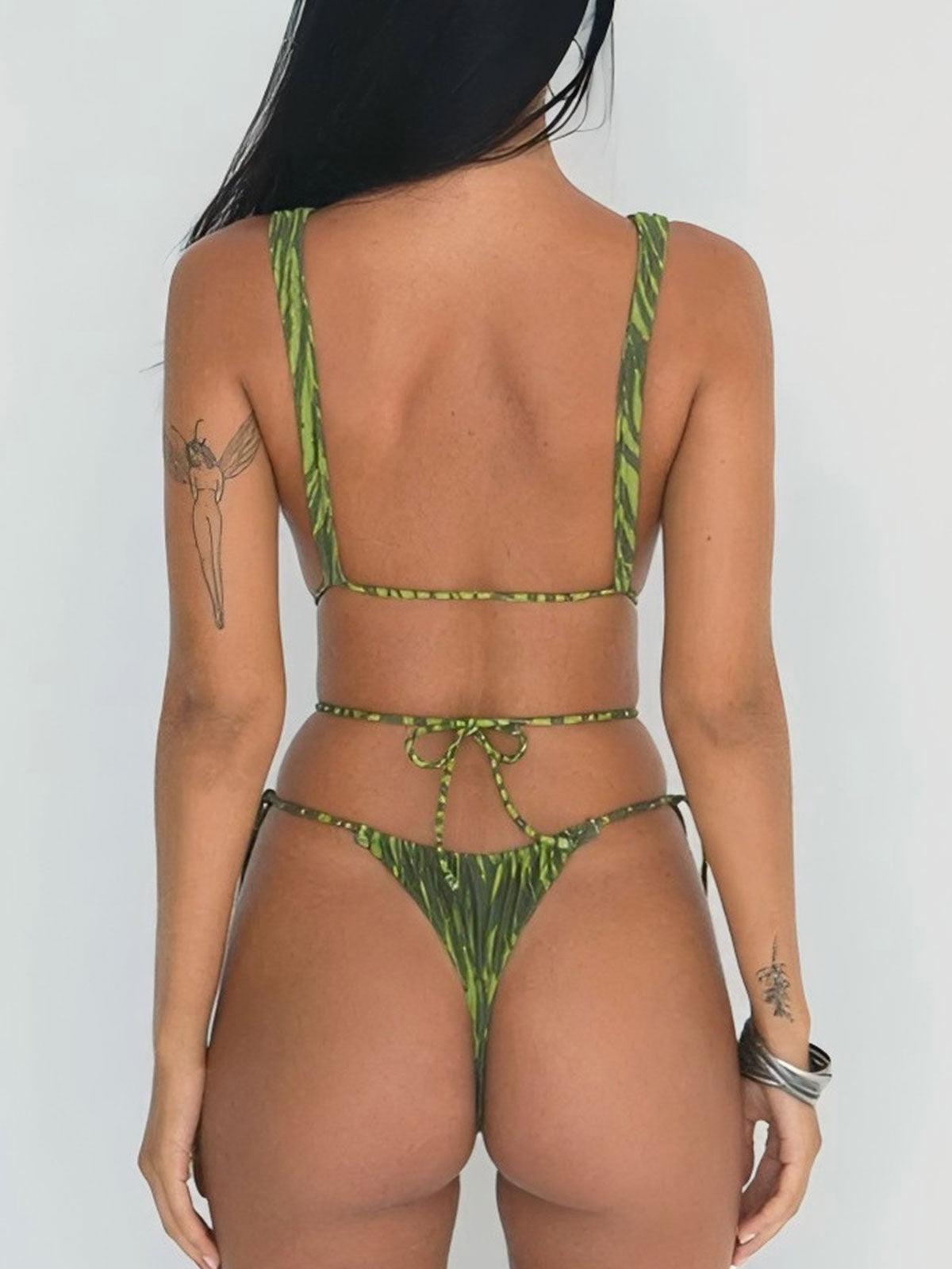 Back view of a woman in a green printed one-piece swimsuit with thin straps and a thong bottom, highlighting the adjustable tie closure.
