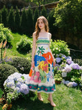 Woman standing in a garden wearing a sleeveless midi dress with a colorful abstract tropical print.
