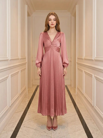 Woman wearing a pink satin midi dress with a twisted knot bodice and pleated skirt, standing in a hallway.