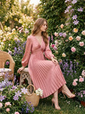 Woman wearing a pink satin midi dress with a twisted knot bodice and pleated skirt, sitting in a garden.