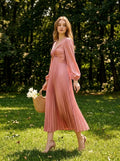 Woman wearing a pink satin midi dress with a twisted knot bodice and pleated skirt, walking in a park.