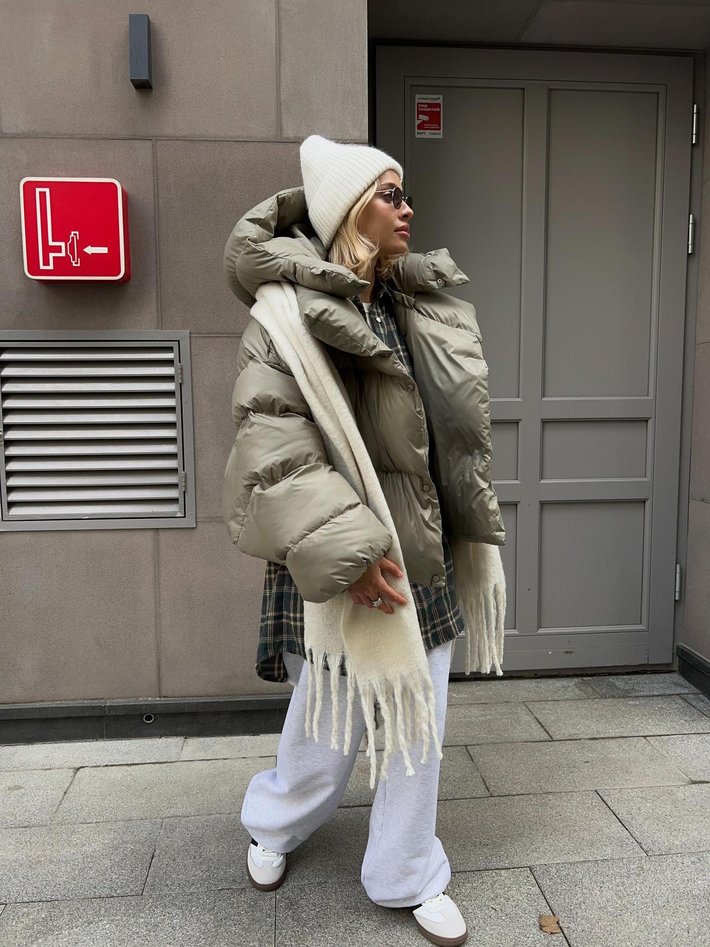 Woman wearing a khaki green oversized puffer jacket with a hood, paired with sweatpants and a scarf.
