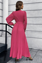 Back view of a woman in a long sleeve pink pleated maxi dress with a v-neck and a self-tie belt.