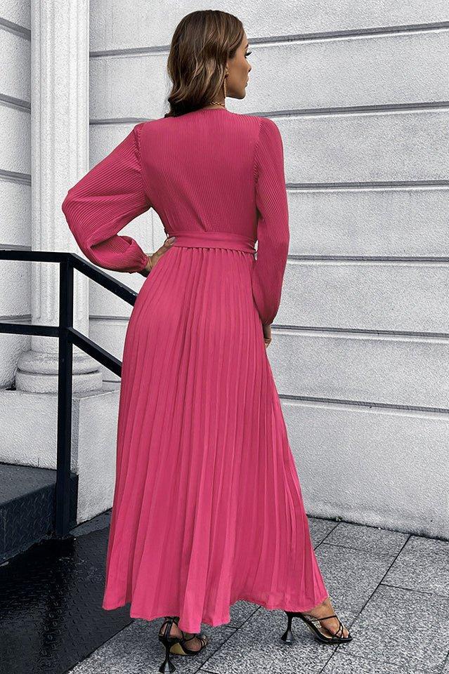 Back view of a woman in a long sleeve pink pleated maxi dress with a v-neck and a self-tie belt.
