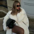 Woman sitting with a black handbag, wearing a long white faux fur coat and sunglasses