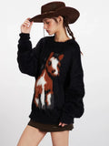 Side view of a woman wearing a black fuzzy knit sweater with a prominent horse graphic, brown cowboy hat, and brown boots, showcasing the relaxed fit.