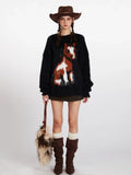 Woman wearing a black fuzzy knit sweater with a brown and white horse graphic on the front, paired with a brown cowboy hat, olive green shorts, white socks, and brown boots.