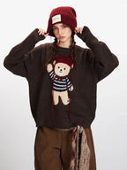 Woman adjusting her red beanie while wearing a brown knit sweater with a teddy bear graphic and brown pants.
