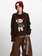 Front view of a woman wearing a brown knit sweater with a teddy bear graphic, brown pants, and a red beanie, showing the graphic detail.