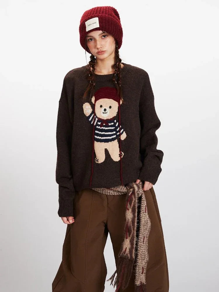 Front view of a woman wearing a brown knit sweater with a teddy bear graphic, brown pants, and a red beanie, showing the graphic detail.