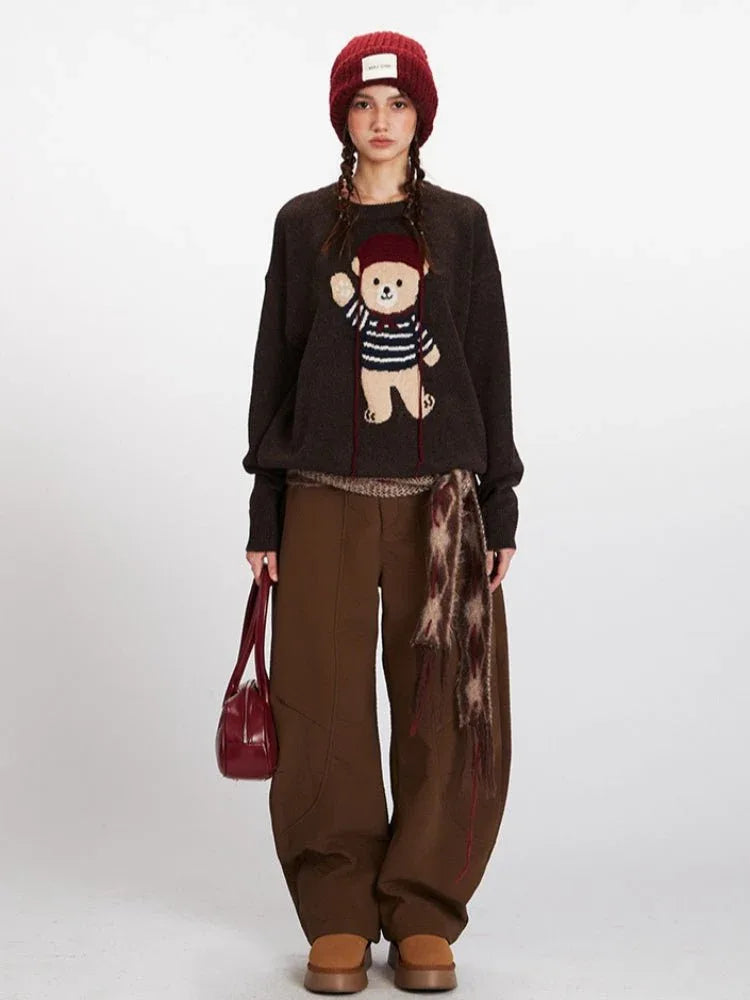 Woman wearing a brown knit sweater with a teddy bear graphic, brown pants, and a red beanie.