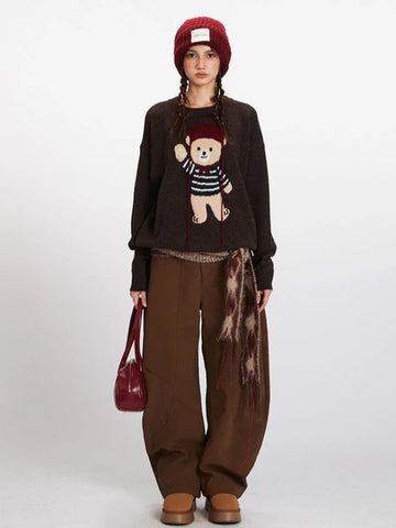 Woman wearing a brown knit sweater with a teddy bear graphic, brown pants, and a red beanie.