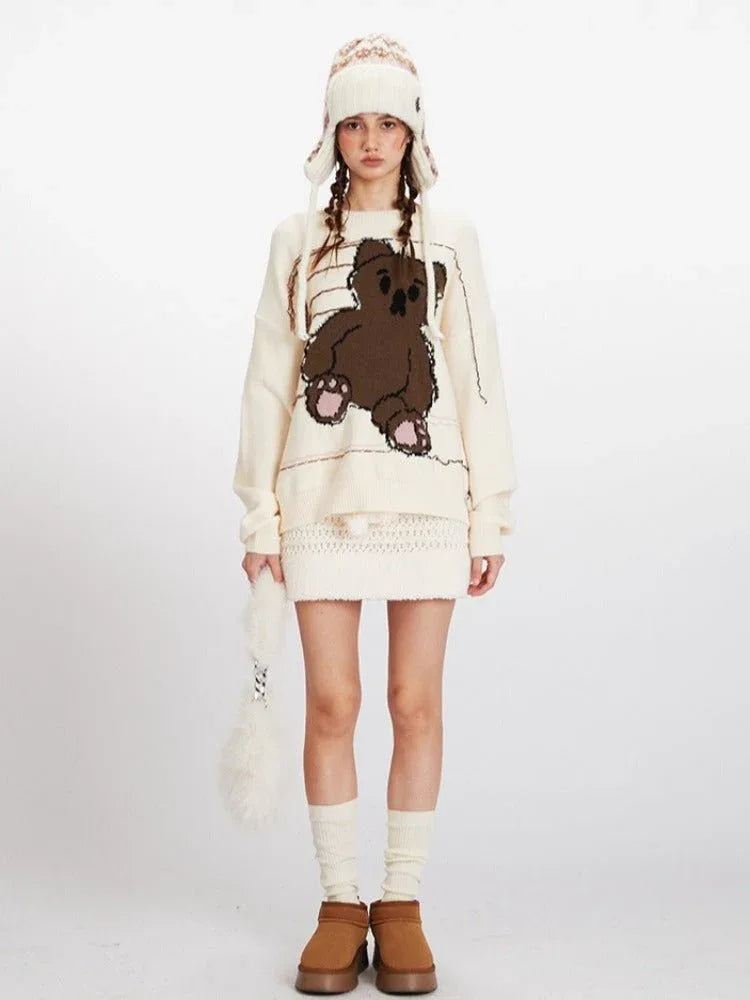 Woman wearing a cream knit sweater with a large brown teddy bear graphic on the front, paired with a cream textured skirt and a patterned beanie.