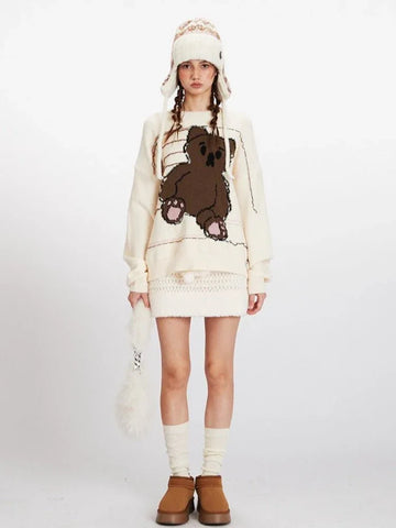 Woman wearing a cream knit sweater with a large brown teddy bear graphic on the front, paired with a cream textured skirt and a patterned beanie.