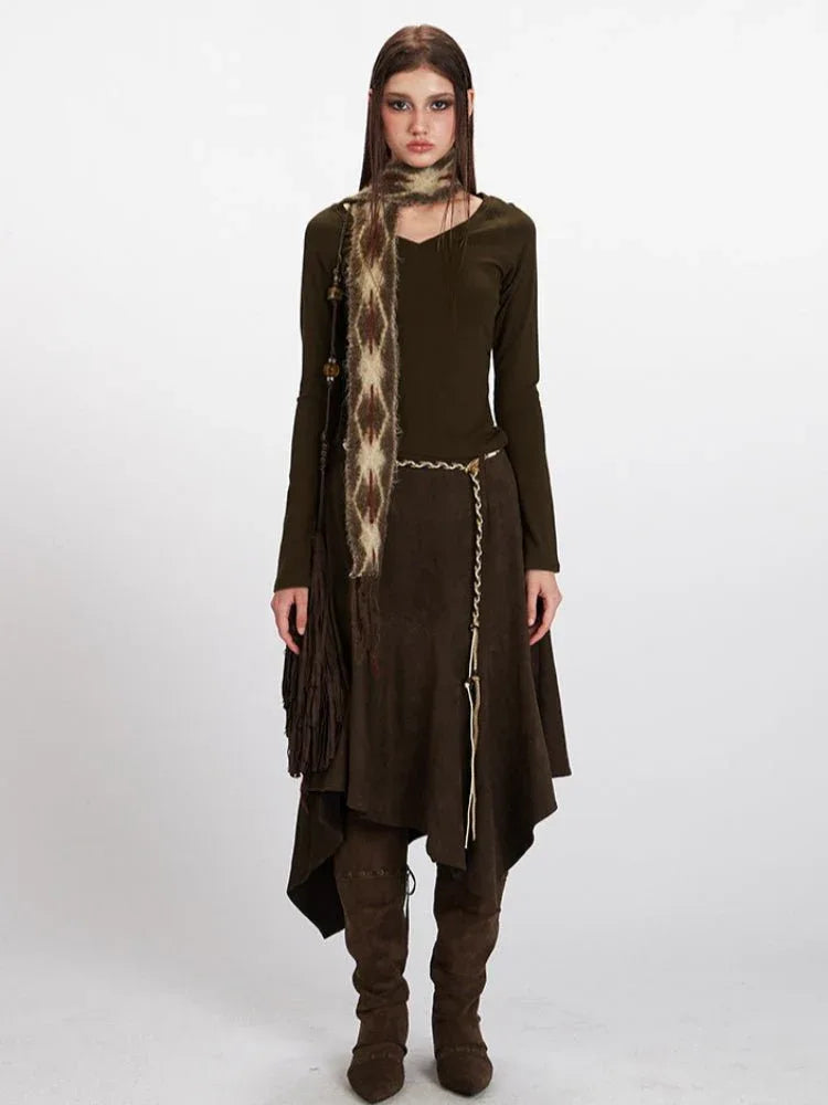 Woman wearing a long sleeve olive green top, a brown asymmetrical hem suede skirt, a patterned scarf, a gold chain belt, and brown suede boots.