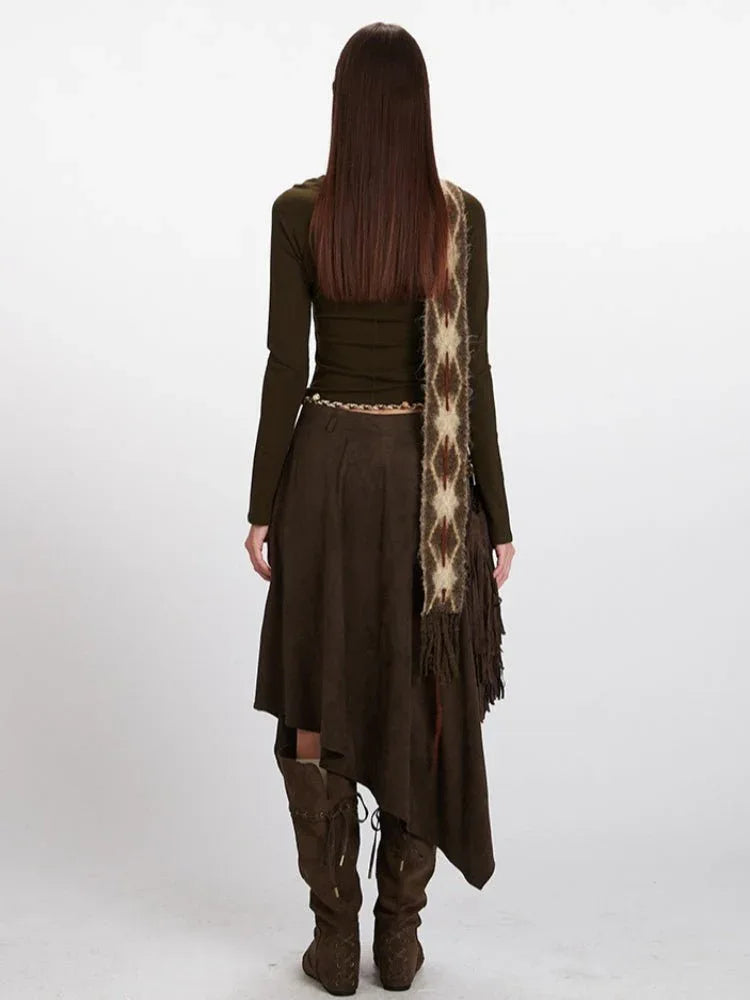 Woman wearing a long sleeve olive green top, a brown asymmetrical hem suede skirt, a patterned scarf, a gold chain belt, and brown suede boots.