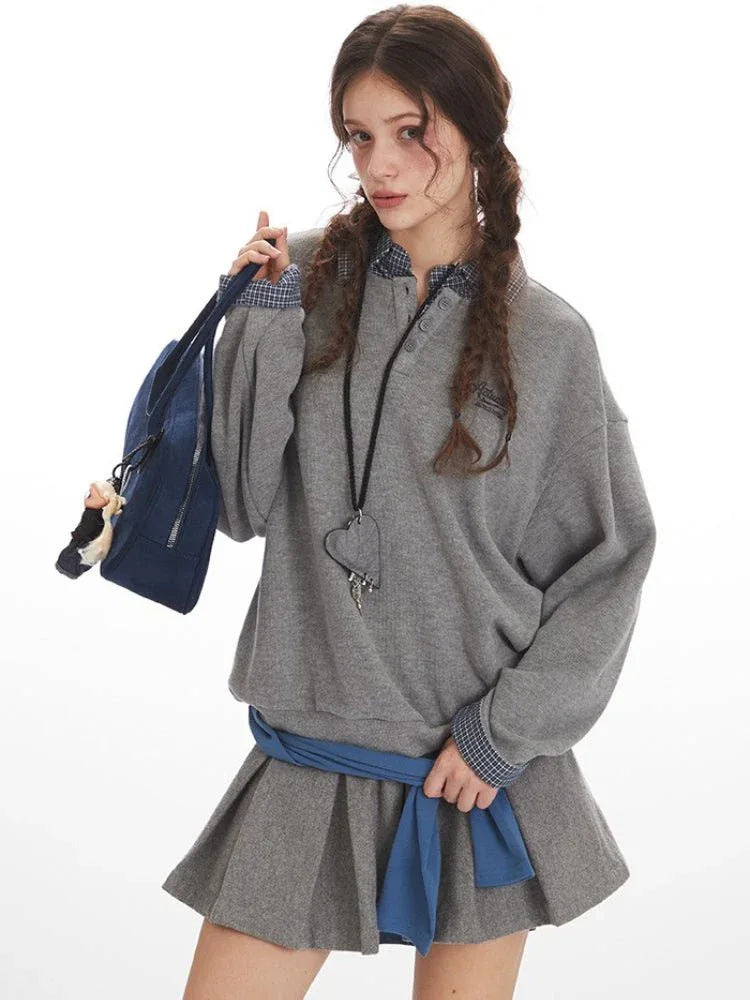 Close-up of a woman wearing a grey oversized sweatshirt over a plaid collared shirt, showing the layered detail and embroidered logo.