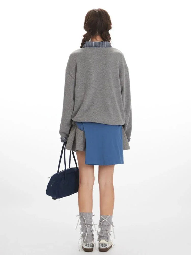 Woman wearing a grey oversized sweatshirt over a plaid collared shirt and a grey pleated skirt, cinched with a blue sash, holding a navy blue duffel bag.
