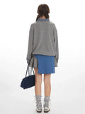 Back view of a woman wearing a grey oversized sweatshirt over a plaid collared shirt and a grey pleated skirt, cinched with a blue sash, holding a navy blue duffel bag.
