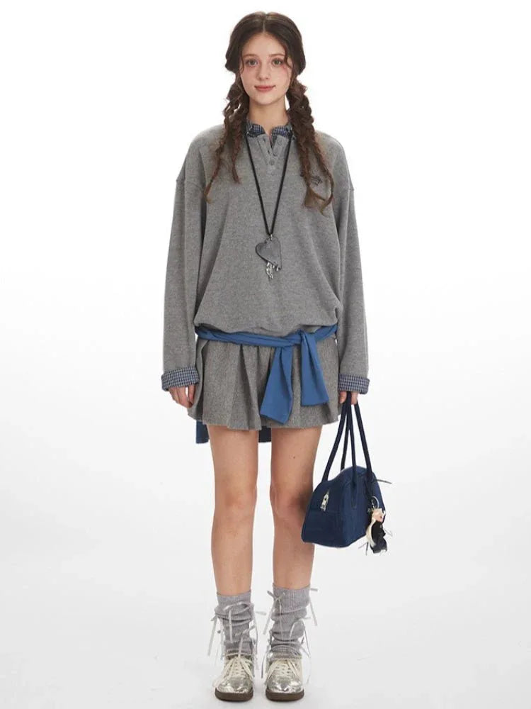 Woman wearing a grey oversized sweatshirt over a plaid collared shirt and a grey pleated skirt, cinched with a blue sash, holding a navy blue duffel bag.