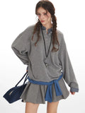 Woman posing with a grey oversized sweatshirt over a plaid collared shirt and a grey pleated skirt, holding a navy blue duffel bag.