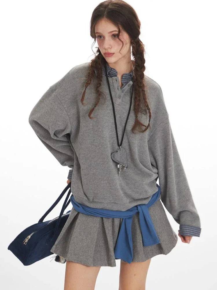 Woman posing with a grey oversized sweatshirt over a plaid collared shirt and a grey pleated skirt, holding a navy blue duffel bag.