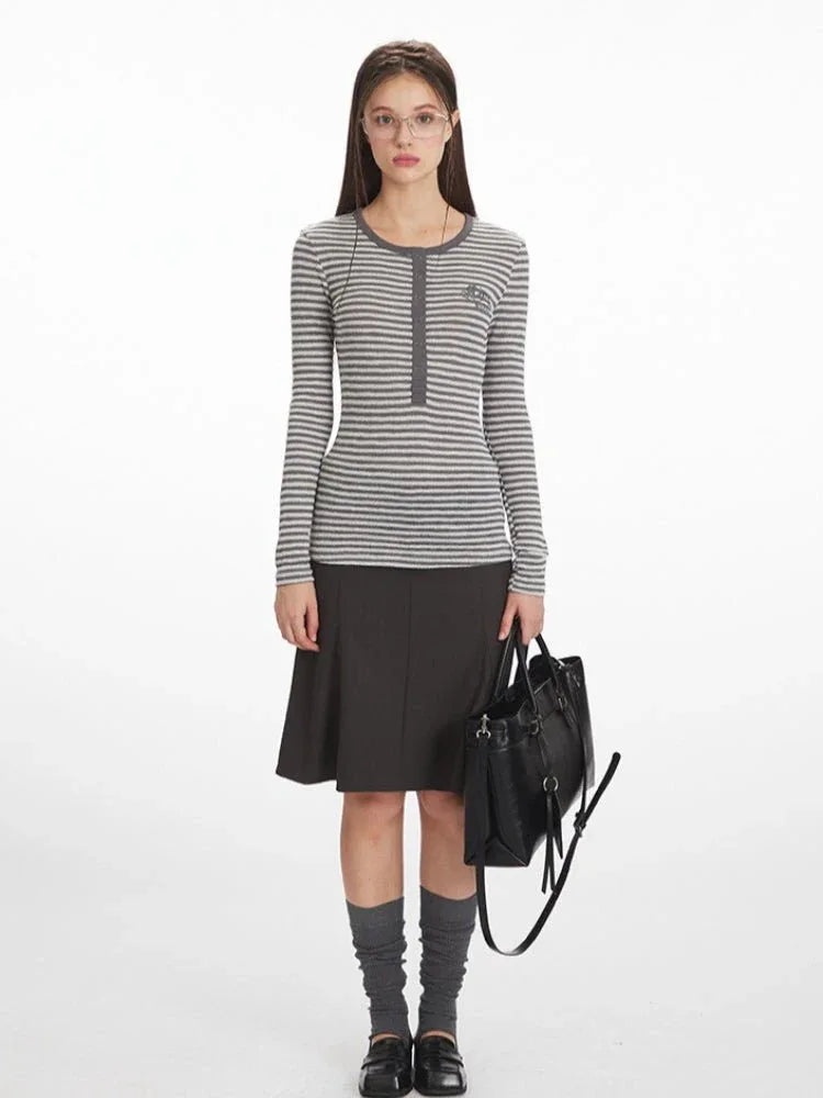 Woman wearing a grey and white striped long-sleeve top with a button-up placket and embroidered detail, paired with a dark grey A-line skirt, grey knee-high socks, and black loafers.