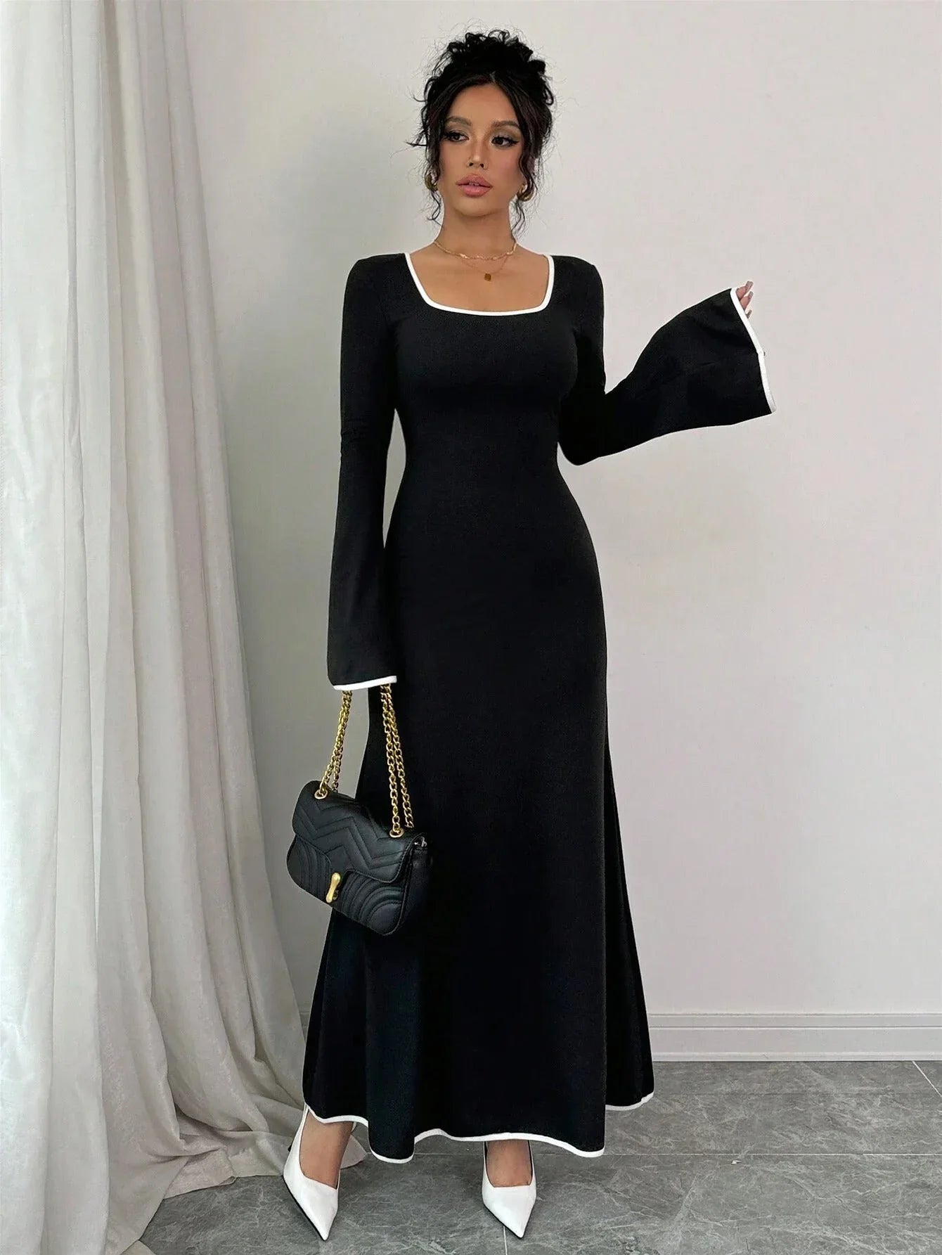 Woman holding a black handbag and wearing a black maxi dress with white contrast piping and bell sleeves.