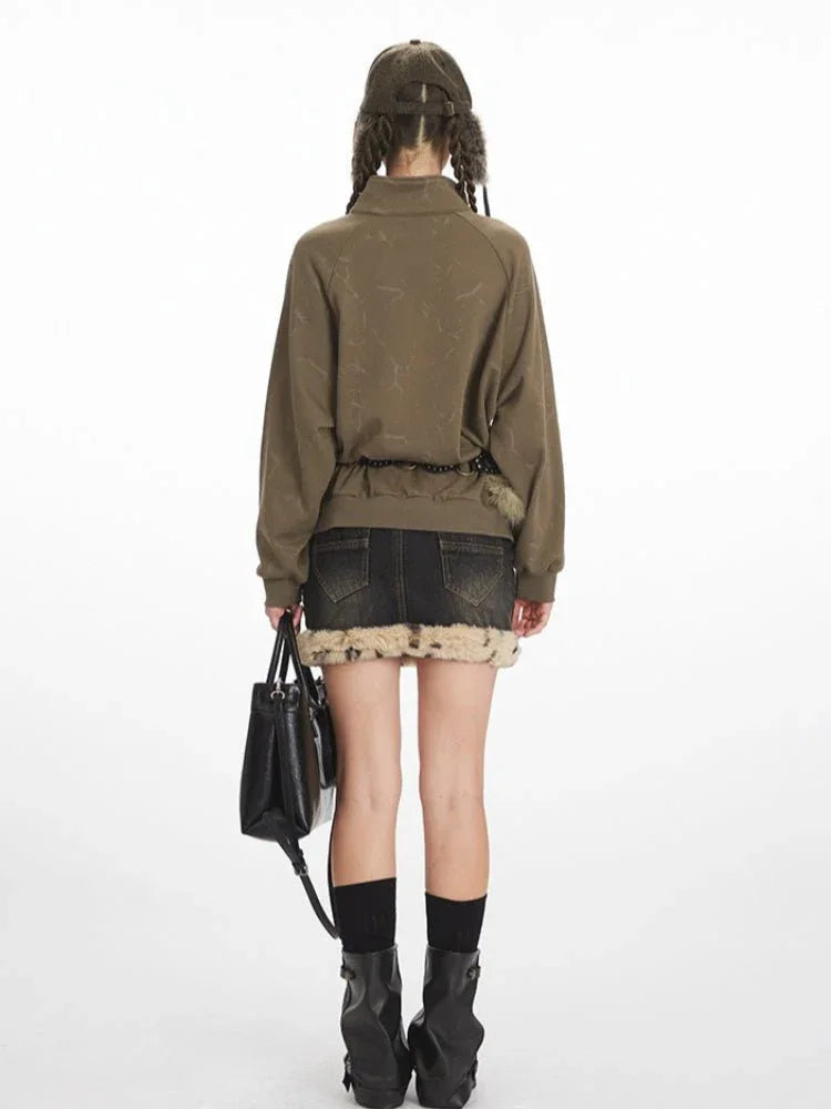 Woman wearing a green graphic print sweatshirt with a collared button placket, black denim skirt with fur trim, black boots, and a black handbag.