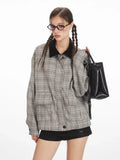 Woman wearing a grey plaid bomber jacket with a black collar, black shorts, black knee-high socks, and black shoes, holding a black handbag and adjusting her glasses.