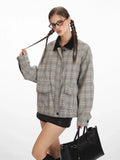 Woman wearing a grey plaid bomber jacket with a black collar, black shorts, black knee-high socks, and black shoes, holding a black handbag and touching her braided hair.