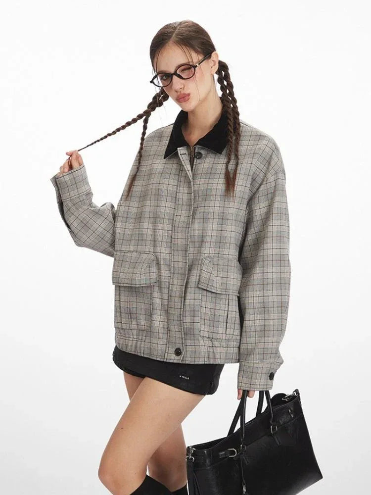Woman wearing a grey plaid bomber jacket with a black collar, black shorts, black knee-high socks, and black shoes, holding a black handbag and touching her braided hair.