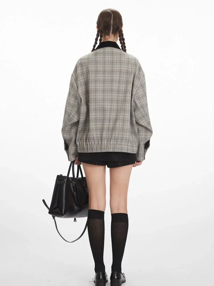 Woman wearing a grey plaid bomber jacket with a black collar, black shorts, black knee-high socks, and black shoes, holding a black handbag.